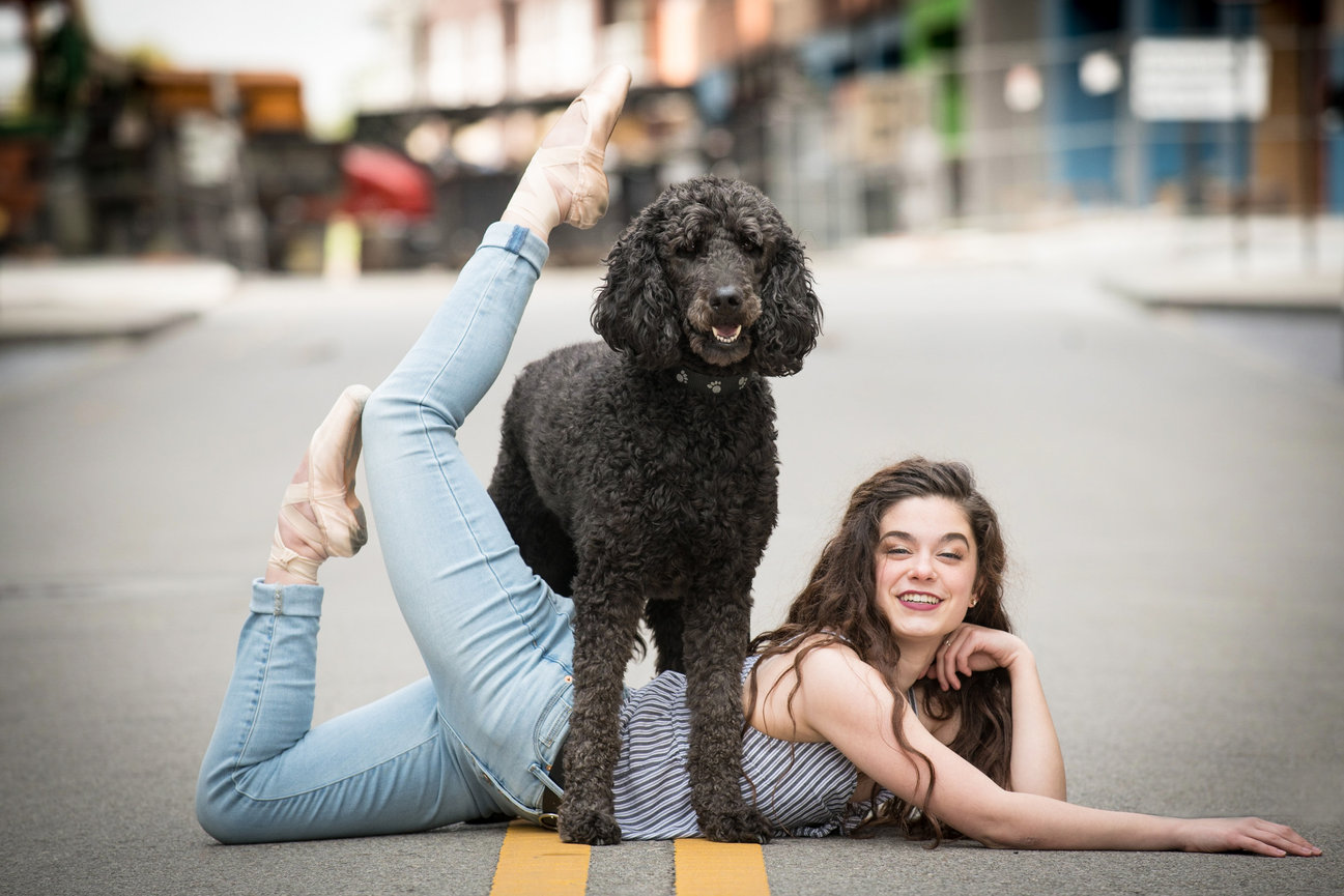 Ballet dancer posing with a black dog on a city street, showcasing flexibility and playfulness.