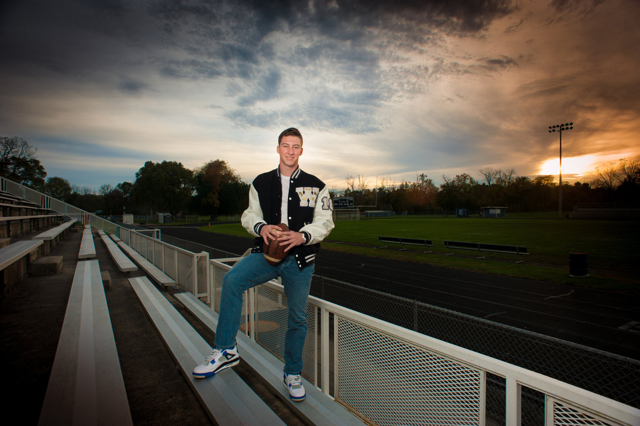 High school senior in a letterman jacket poses with a football on bleachers at sunset.