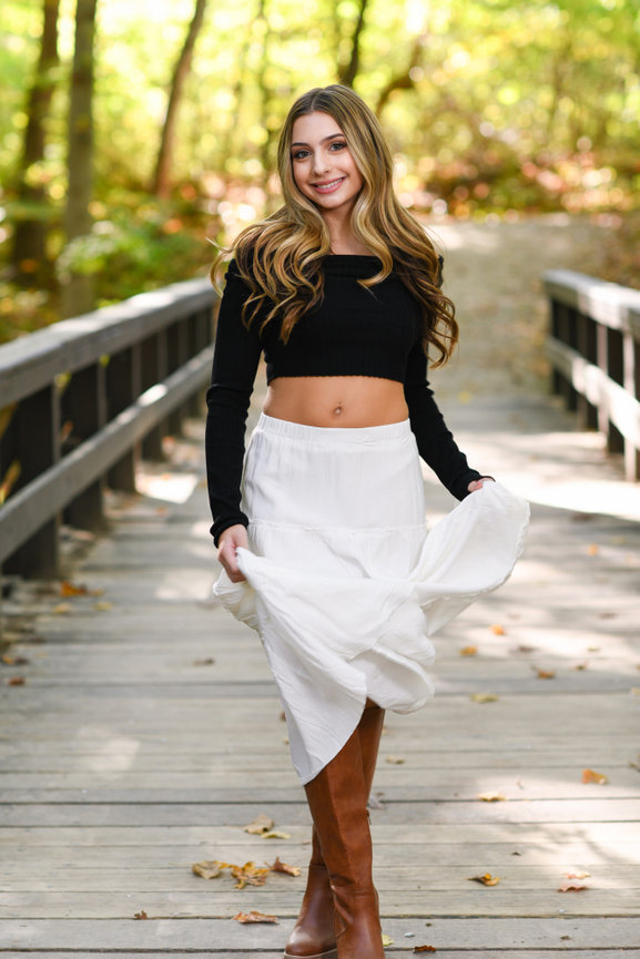 A teenage girl with a white skirt and black crop top walking on a rustic bridge in the forest.