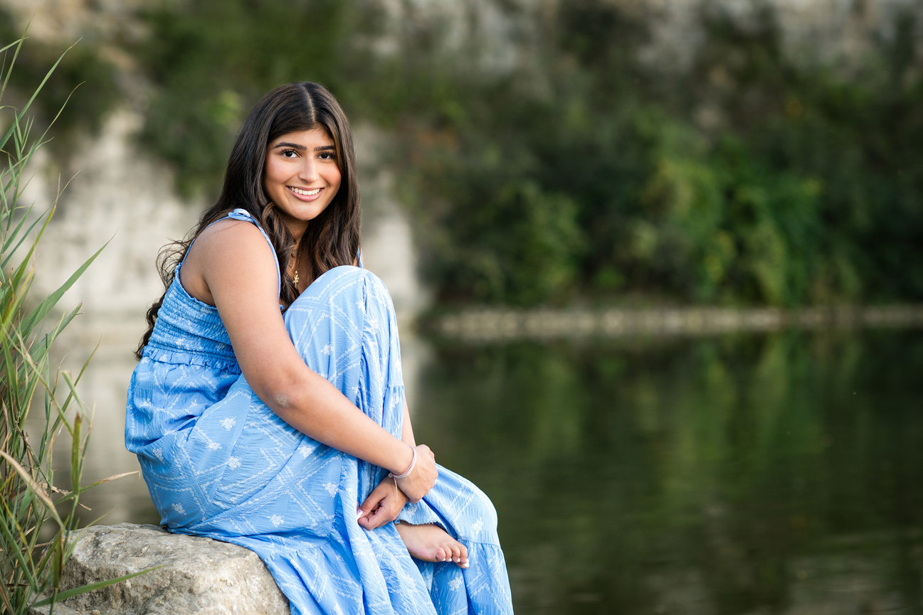 Teen girl in blue dress sitting on a rock by a calm lake, smiling, with greenery in the background. Senior pictures Columbus Ohio.