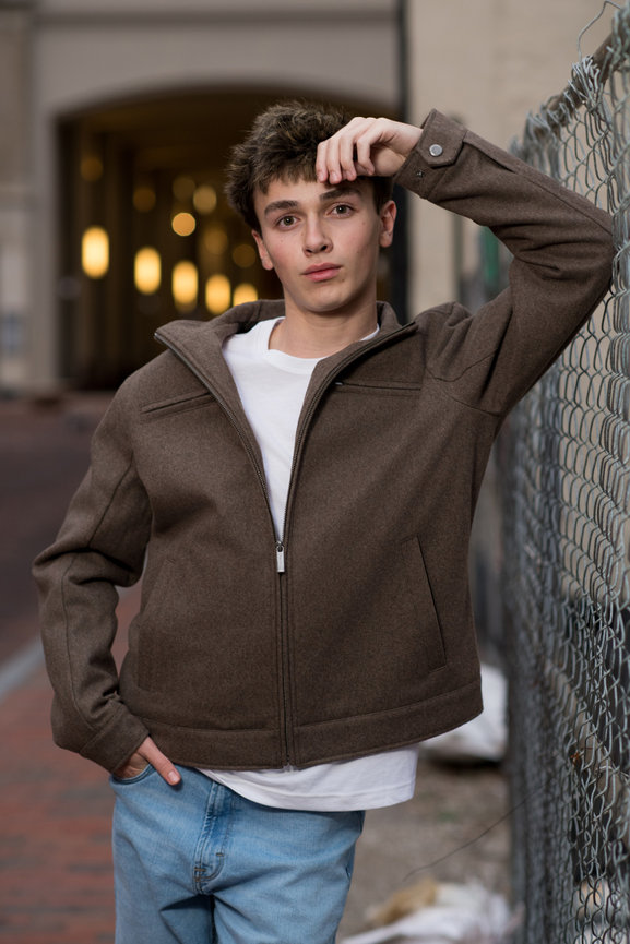 Teenage boy in a brown jacket and jeans, leaning on a chain-link fence with an urban background. Senior pictures in Columbus Ohio