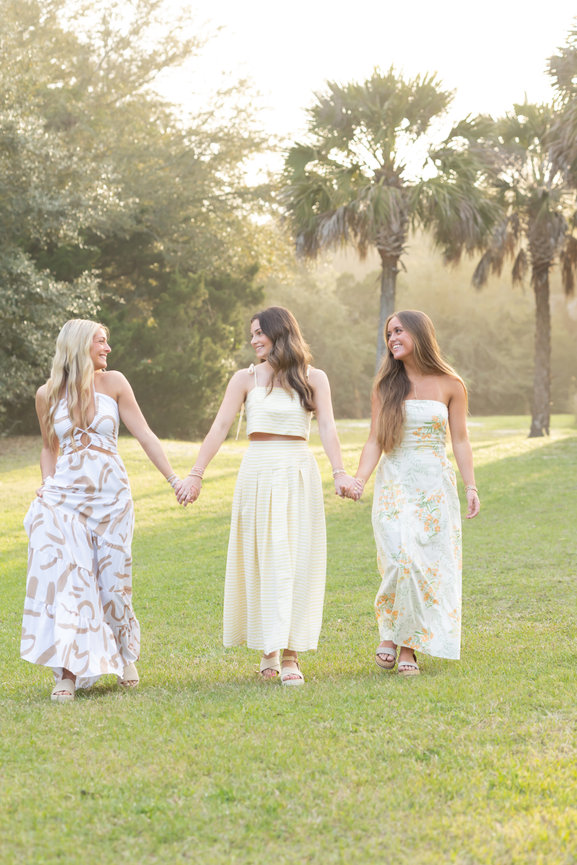 Three women in summer dresses holding hands and walking on grass with palm trees behind them