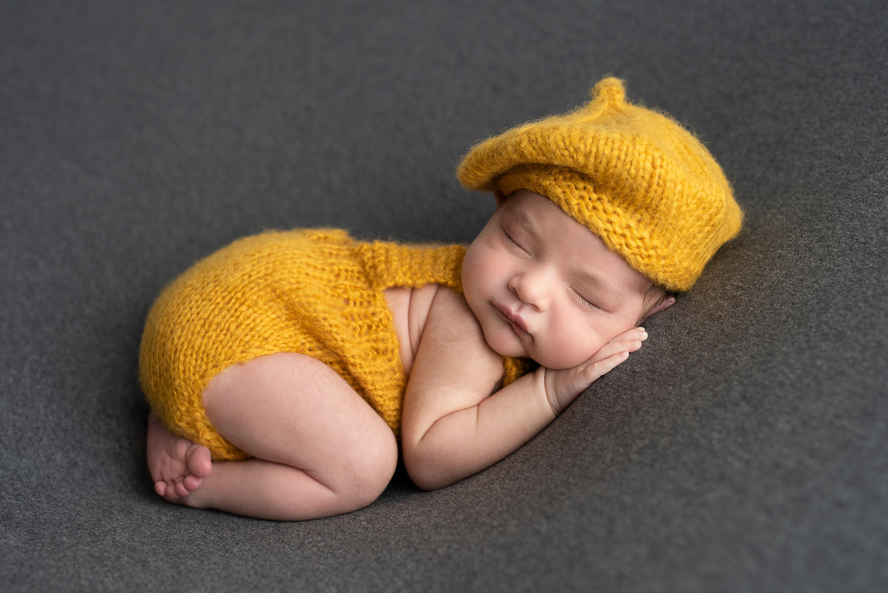 A sleeping baby wearing a matching yellow knit outfit and hat is curled up on a soft, dark gray surface, bringing an aura of peace and coziness to the Carpentersville home in Illinois.