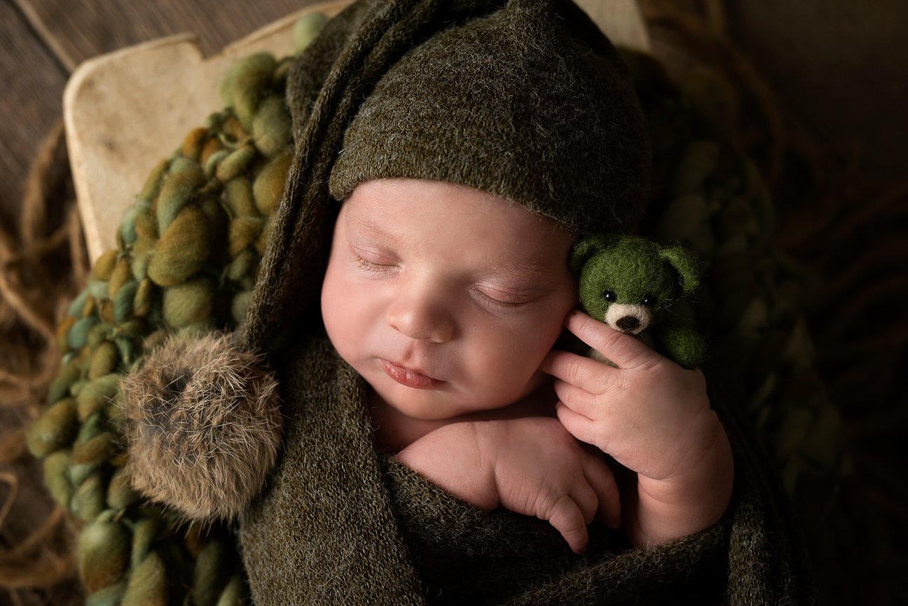 A sleeping baby wrapped in a cozy green blanket, wearing a matching knitted hat with a pom-pom, cuddles a tiny green teddy bear. The peaceful scene, reminiscent of autumn in Hoffman Estates, unfolds on a textured surface with brown and green tones.